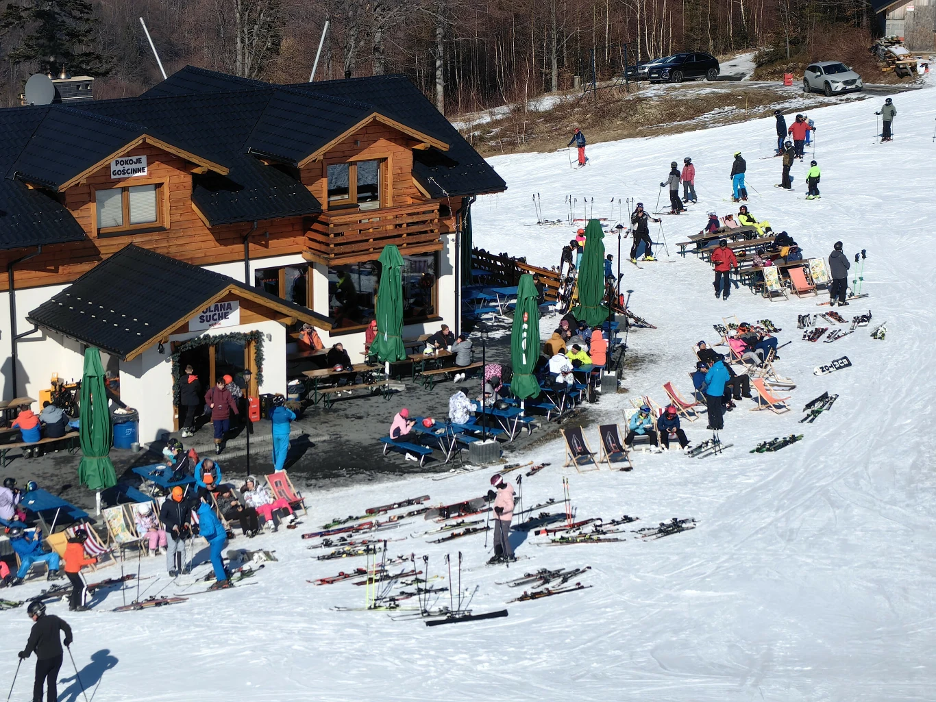 Drewniany budynek schroniska górskiego otoczony przez narciarzy wypoczywających na leżakach i korzystających z uroków stoku, liczne narty ustawione wokół, słoneczna pogoda i zimowy krajobraz.