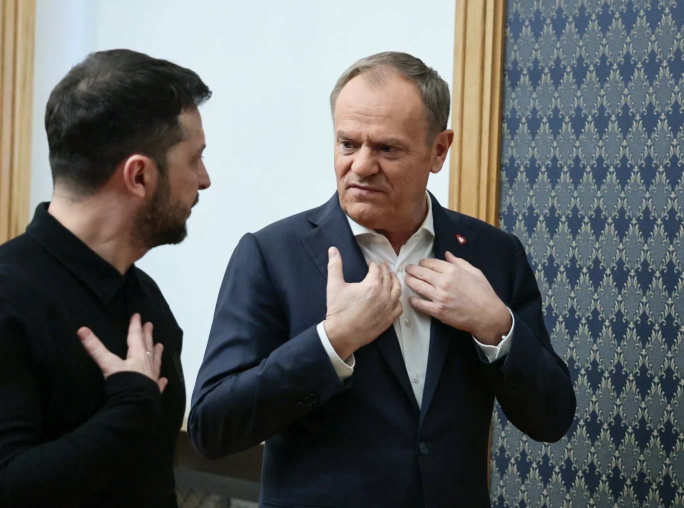Zmiana przepisów w sprawie Ukraińców. Poznaliśmy plany rządu Dwóch mężczyzn prowadzi rozmowę, jeden z nich gestykuluje, obaj stoją w eleganckim otoczeniu z ozdobną tapetą i drewnianymi framugami.