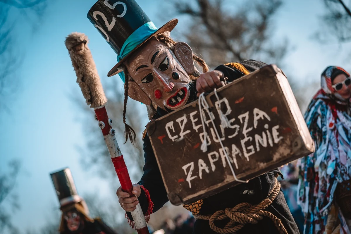 Współcześnie Dziady mają charakter barwnego widowiska Osoba w tradycyjnym stroju ludowym z dużą maską na twarzy, cylindrem z napisem 25 oraz dużą walizką z napisem 'Czego dusza zapragnie', trzymająca ozdobiony kij. W tle inne postacie w przebraniach oraz rozmyte drzewa.