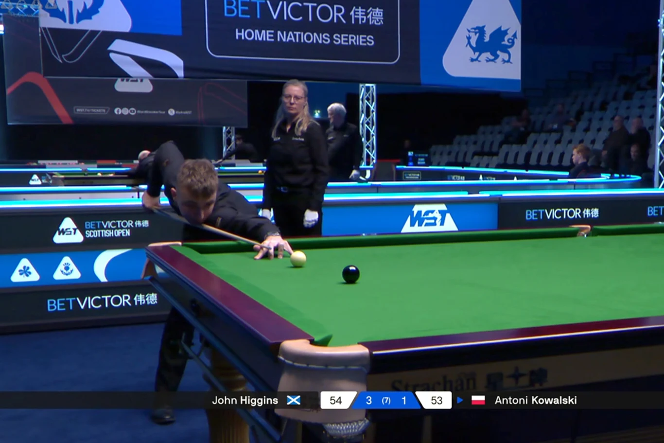 Zawodnik snookera pochylony nad stołem przygotowuje się do uderzenia bili, na stole widoczna pojedyncza bila, w tle sędzia i pustawa widownia, na tablicy wyników nazwiska graczy i aktualny wynik.