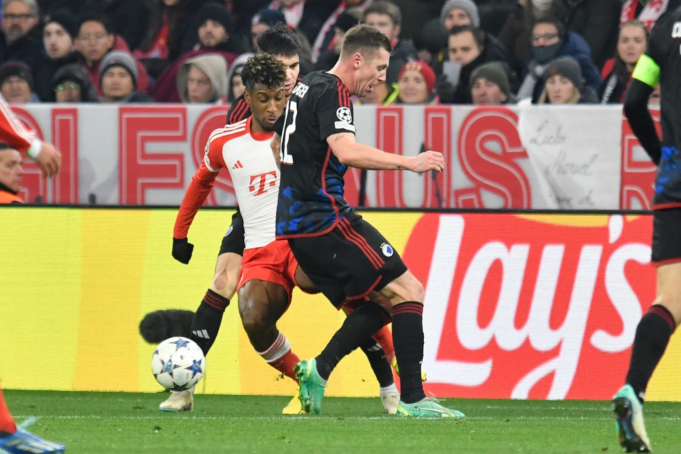 Dwóch piłkarzy walczy o piłkę podczas meczu piłki nożnej na stadionie, otoczeni przez innych zawodników oraz widownię na trybunach.