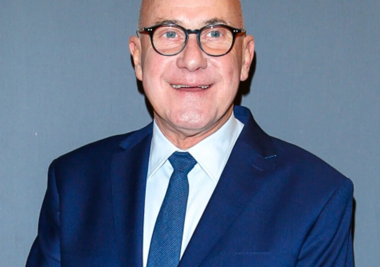 Piotr Machalica Uśmiechnięty mężczyzna w garniturze i krawacie, noszący okulary, stoi na neutralnym tle. Jego postura jest wyprostowana, a wyraz twarzy pogodny.