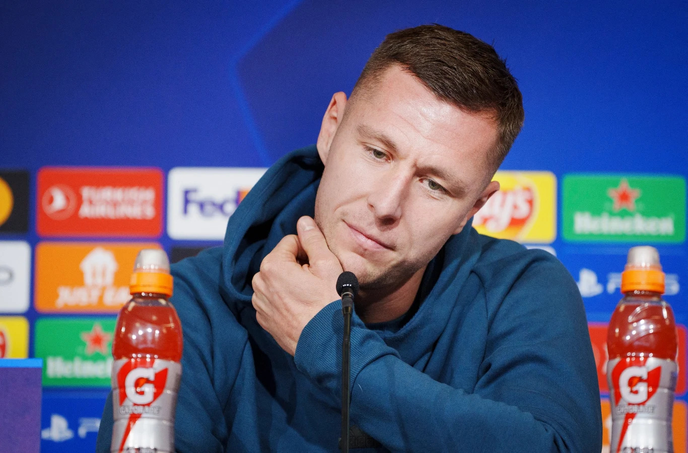 Mężczyzna w niebieskiej bluzie siedzący przy mikrofonie podczas konferencji prasowej, na stole dwa napoje izotoniczne, w tle logotypy sponsorów i organizatorów wydarzenia sportowego.