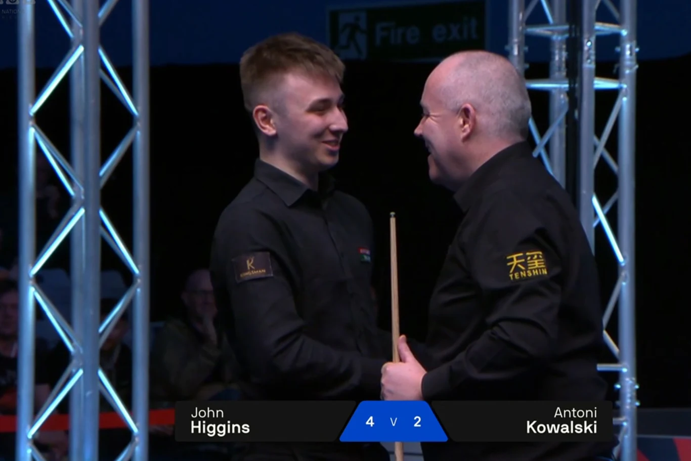 Dwóch snookerzystów w ciemnych koszulach stoi naprzeciw siebie na arenie, rozmawia i uśmiecha się po zakończonej partii, na ekranie widoczny wynik meczu z nazwiskami zawodników.