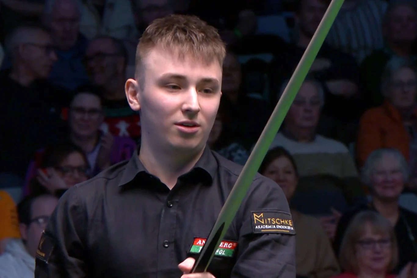 Młody mężczyzna w ciemnej koszuli trzyma kij bilardowy podczas rozgrywki snookera, w tle widoczna publiczność uważnie obserwująca mecz.