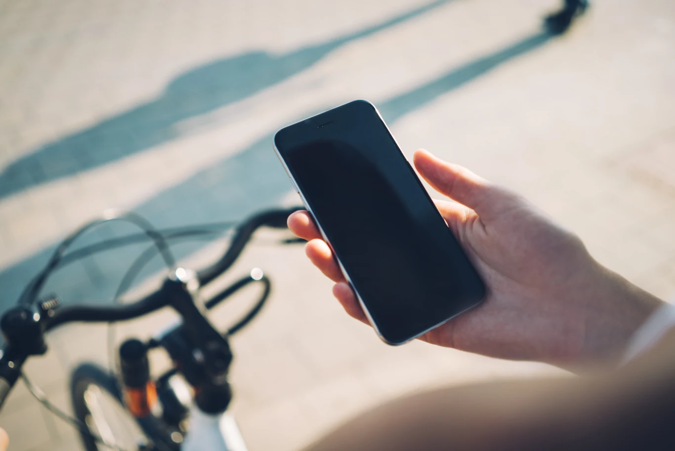 Rejestracja roweru w systemie BIN kosztuje jednorazowo 129 zł. Taka kwota może zniechęcić wielu rowerzystów Dłoń trzymająca smartfona nad kierownicą roweru, w tle rozmyta powierzchnia chodnika i cień.