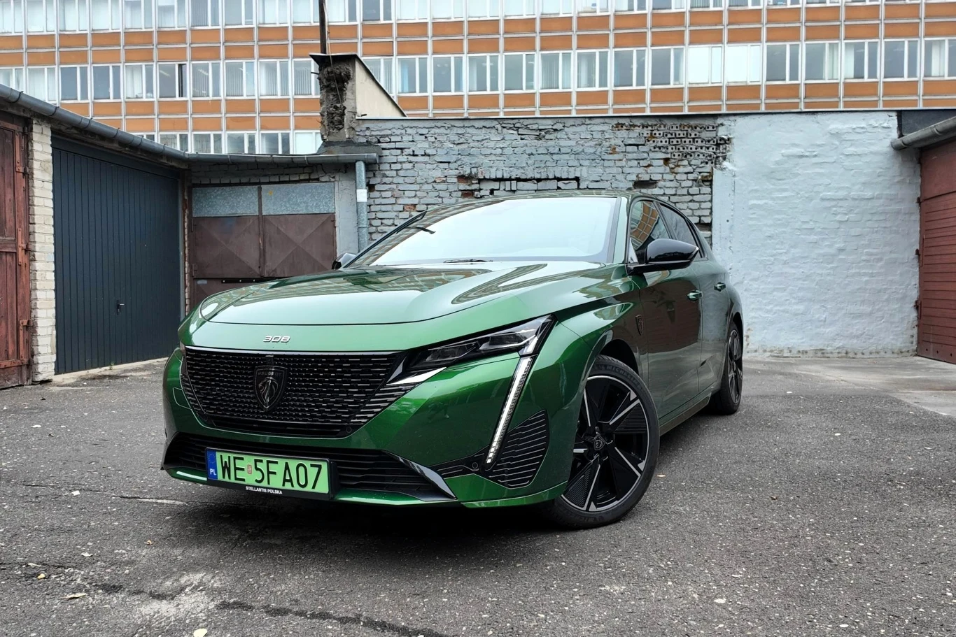 Peugeot E-308 przed modernizacją ciągle jest w ofercie. Czy warto go kupić? Nowoczesny, zielony samochód osobowy marki Peugeot z czarnymi felgami stoi na asfaltowym podjeździe przed garażami, w tle ceglana i betonowa zabudowa oraz przeszklony budynek.
