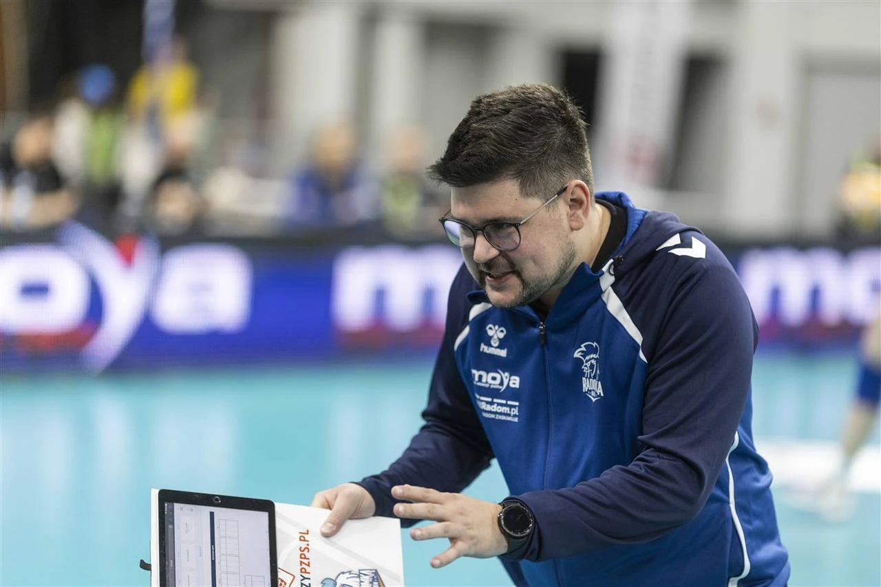Trener drużyny siatkarskiej w sportowym stroju koncentruje się nad notatkami i ekranem podczas przerwy w meczu, w tle widoczna zamazana hala sportowa oraz fragmenty boiska.