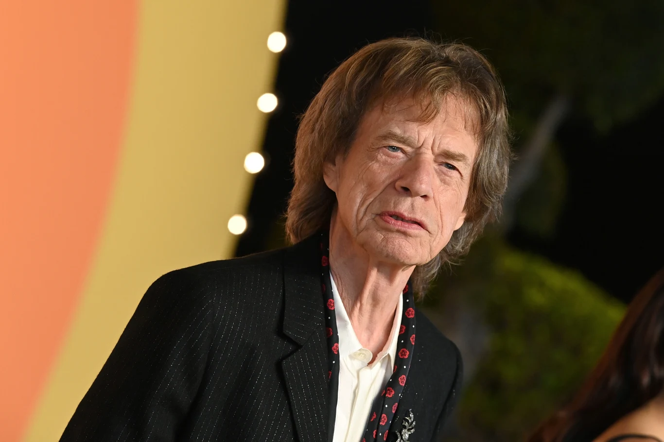 Mick Jagger jest wokalistą The Rolling Stones Mężczyzna w średnim wieku w eleganckiej czarnej marynarce i jasnej koszuli, stojący na tle z rozmytymi światłami i żółtym fragmentem ściany, w tle widoczne zarysy roślinności.