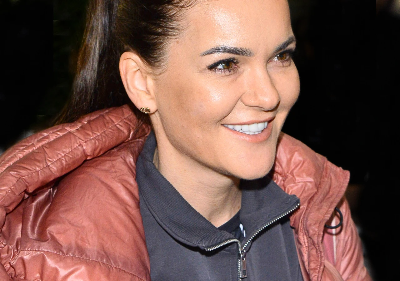 Agnieszka Radwańska Kobieta z długimi ciemnymi włosami spiętymi w kucyk, uśmiechnięta, ubrana w różową kurtkę i szarą bluzę, z widocznym naturalnym makijażem.