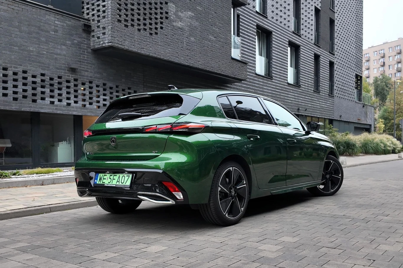 Peugeot E-308 hatchback przed modernizacją rozpędza się do 100 km/h w 9,8 s. Zielony samochód osobowy marki Peugeot stoi zaparkowany na ulicy obok nowoczesnego budynku z ciemnej cegły, otoczonego roślinnością i innymi budynkami w tle.