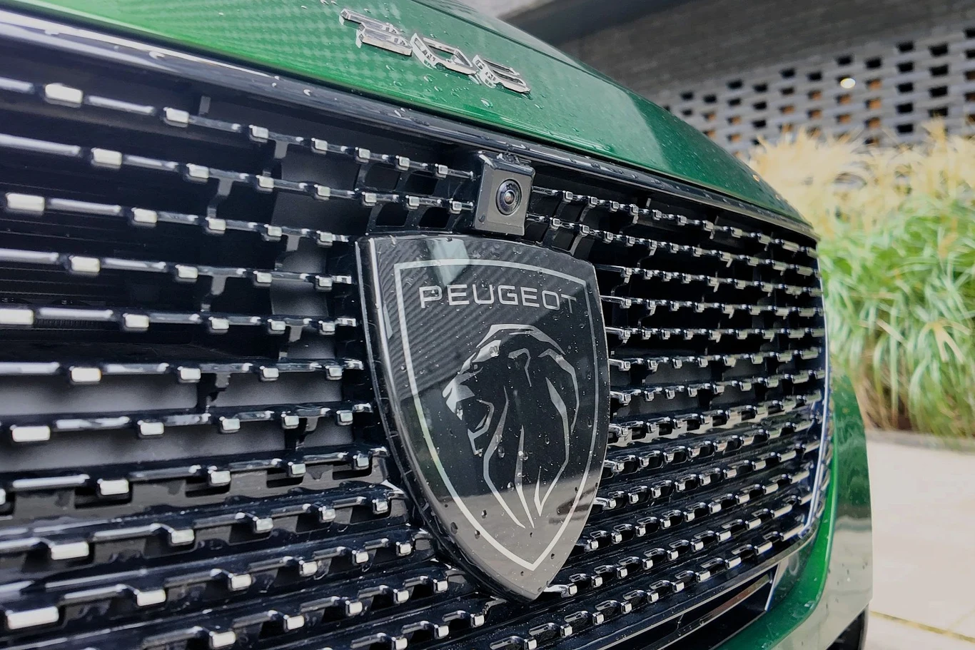 Osoby chcące skorzystać z programu NaszEauto i rozważające wybór Peugeota E-308 mogą śmiało brać pod uwagę wersję przed liftingiem. Duży grill samochodowy z wyraźnym logo marki Peugeot pośrodku, otoczony metalicznymi elementami na zielonym tle maski pojazdu.