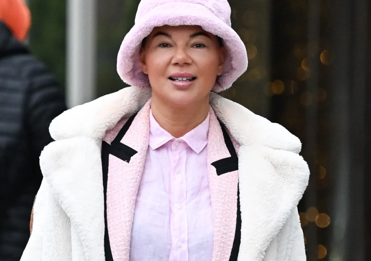 Edyta Górniak pod siedzibą Polsatu Kobieta w różowym kapeluszu i eleganckim płaszczu w jasnych kolorach, ubrana w koszulę z kołnierzykiem, uśmiechnięta, sfotografowana na zewnątrz.