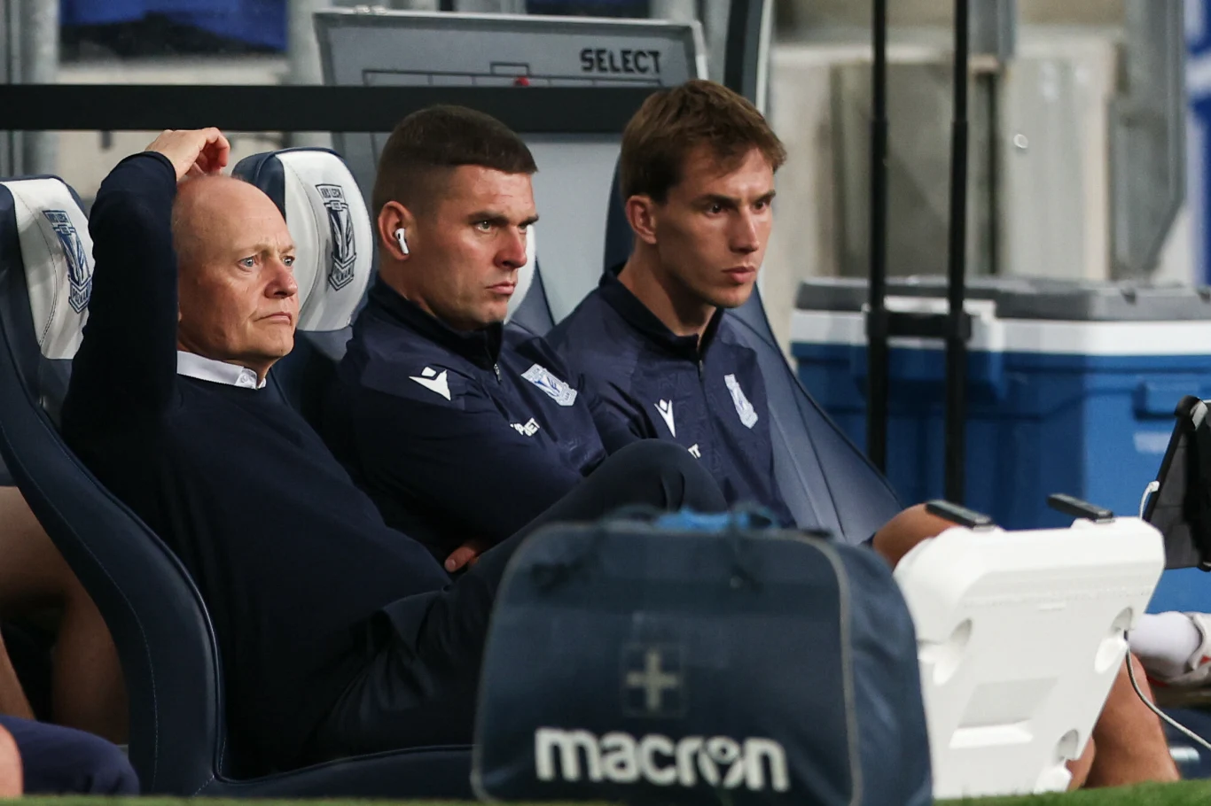 Trzech mężczyzn ubranych w sportowe bluzy siedzi na ławce rezerwowych podczas meczu piłkarskiego, jeden z nich opiera rękę na oparciu fotela, dwoje patrzy uważnie przed siebie, w tle widoczne elementy stadionu i torby sportowe.