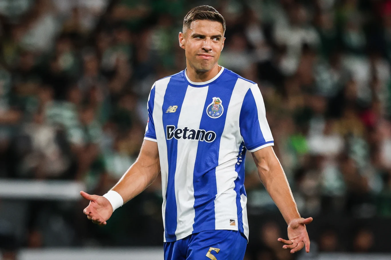 Piłkarz w stroju FC Porto podczas meczu na boisku, wykonujący gest wzruszenia ramionami, w tle rozmazana publiczność stadionu.