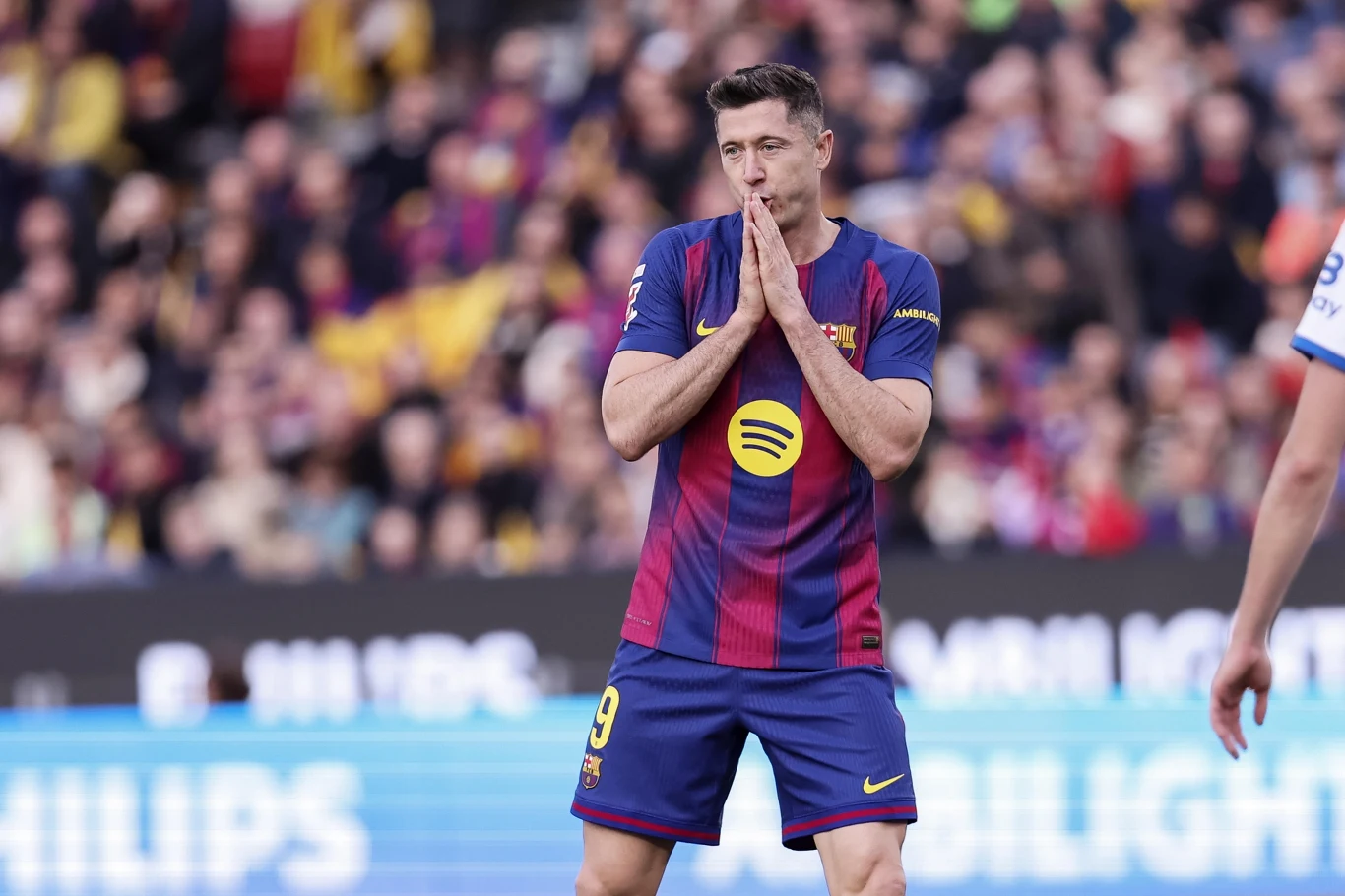 Robert Lewandowski Piłkarz w koszulce FC Barcelony z numerem 9 stoi na murawie, trzymając złożone dłonie przy twarzy w geście rozczarowania lub modlitwy. W tle rozmyty tłum kibiców na stadionie.