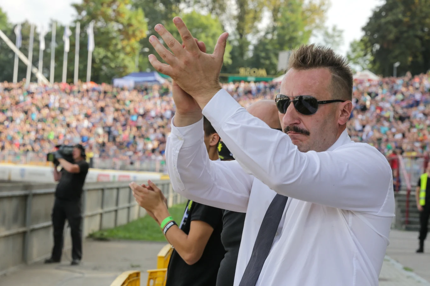 Mężczyzna w białej koszuli i okularach przeciwsłonecznych klaszcze w tłumie podczas wydarzenia sportowego, wokół widoczna publiczność i kilka innych osób, tło stanowią trybuny pełne ludzi.