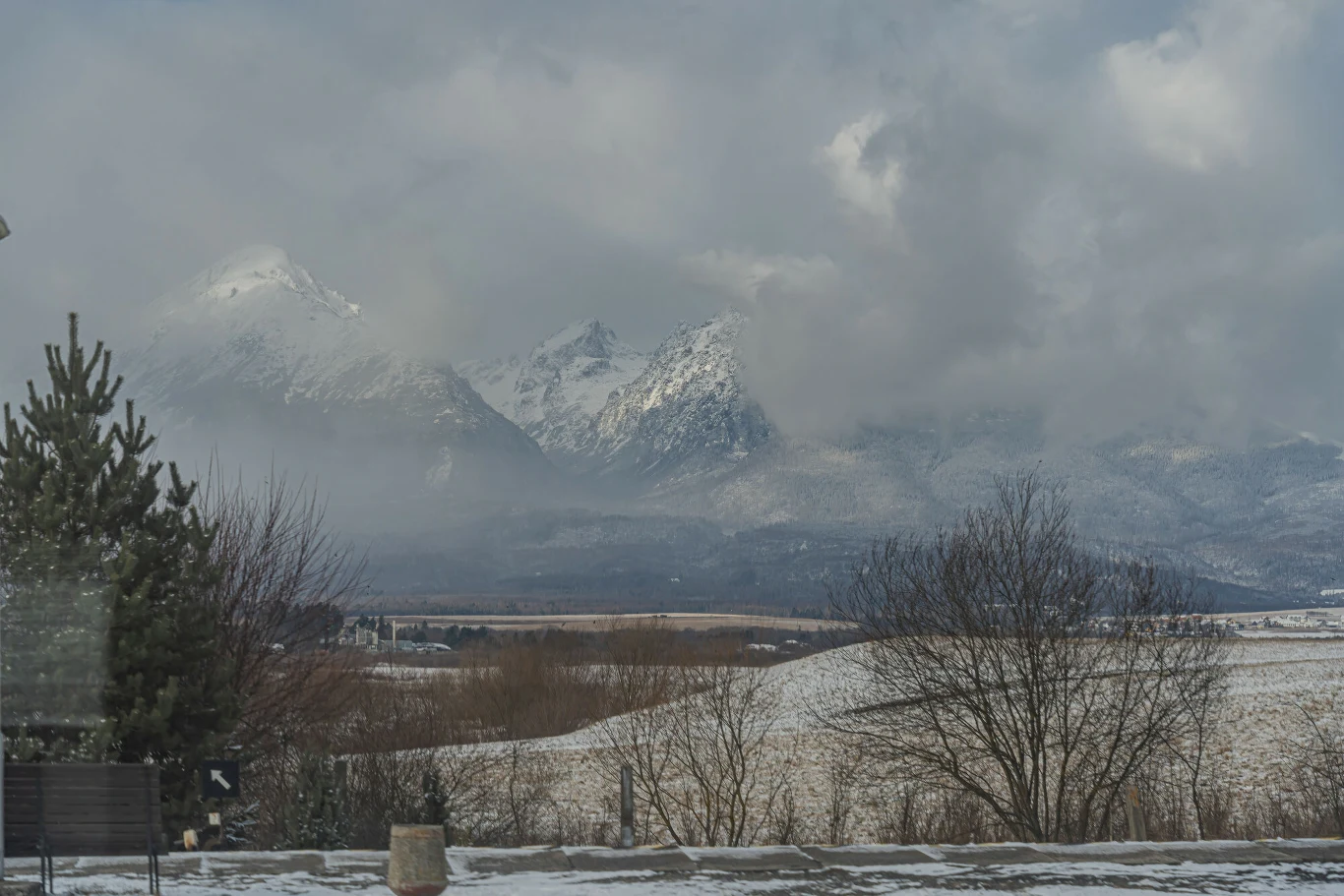 Pokryte śniegiem szczyty górskie otoczone chmurami w tle, na pierwszym planie pola i drzewa bez liści, częściowo przykryte cienką warstwą śniegu, w oddali widać fragment zabudowań.