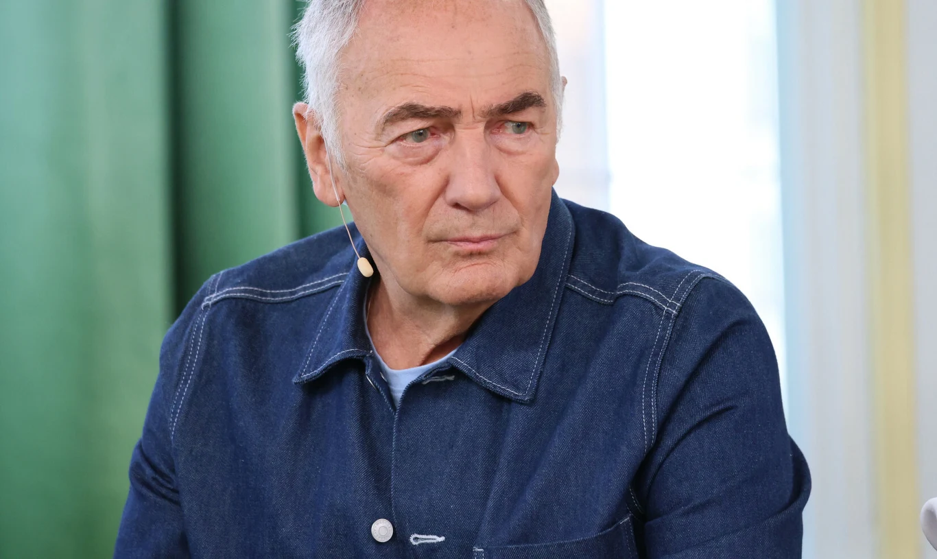 Karol Strasburger Starszy mężczyzna o siwych włosach ubrany w granatową kurtkę z dżinsu, siedzi we wnętrzu ze światłem dziennym i zieloną zasłoną w tle, z mikrofonem przypiętym do kołnierza.