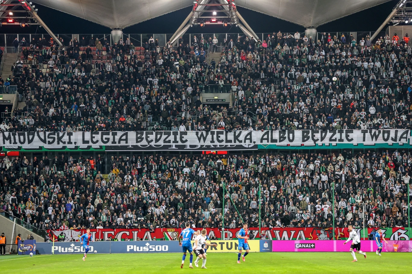 Kibice Legii Warszawa mają już serdecznie dość mijającej rundy Stadion piłkarski wypełniony kibicami Legii Warszawa, którzy trzymają długi transparent z napisem skierowanym do właściciela klubu. Na murawie trwa mecz piłki nożnej, widoczni są zawodnicy obu drużyn.