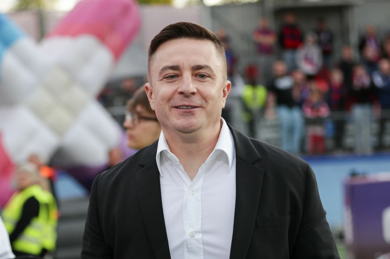 Mężczyzna w białej koszuli i czarnej marynarce stoi na tle ludzi oraz dużej dmuchanej instalacji w kolorze różowym i niebieskim, w otwartej przestrzeni, prawdopodobnie podczas wydarzenia sportowego lub kulturalnego.