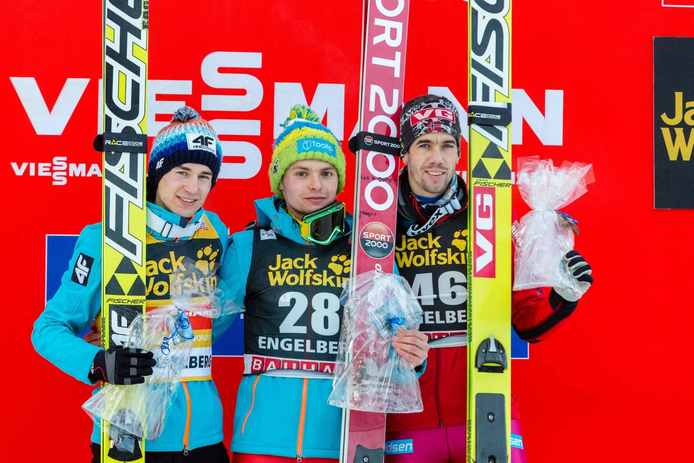Jan Ziobro w Engelbergu wygrał jedyny konkurs Pucharu Świata. Na podium razem z Kamilem Stochem i Andersem Bardalem Trzech sportowców w strojach narciarskich stoi na podium, każdy trzyma narty i upominki, w tle znajdują się logotypy sponsorów i intensywnie czerwone tło zawodów.