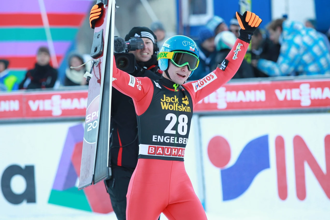 Jan Ziobro Zawodnik w czerwonym stroju narciarskim i kasku z podniesionymi rękami w geście triumfu, trzymający narty, na tle band reklamowych podczas zawodów sportowych.