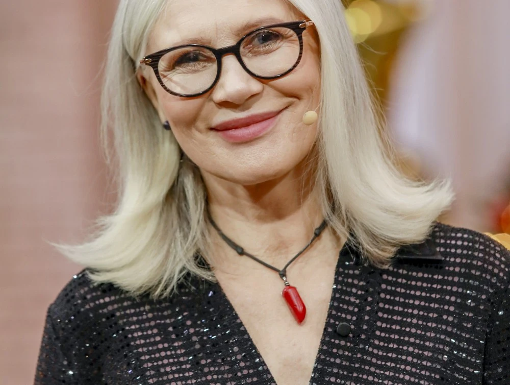 Joanna Dyrkacz znalazła nową miłość Starsza kobieta z jasnymi włosami i okularami, uśmiecha się, mając na sobie czarną, błyszczącą bluzkę oraz naszyjnik z czerwonym wisiorkiem, tło rozmyte i utrzymane w ciepłych kolorach.
