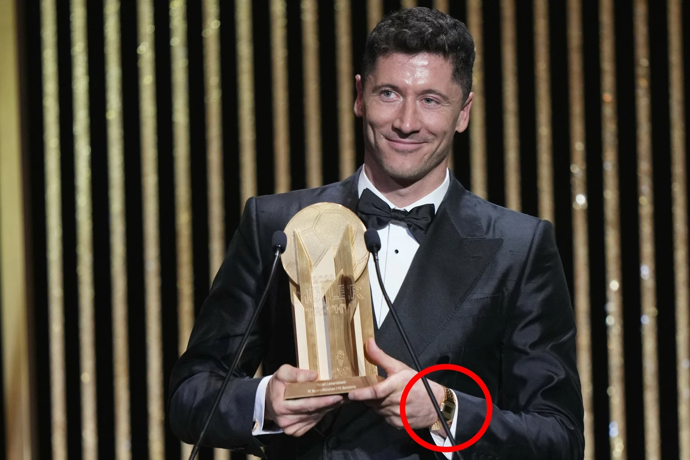 Mężczyzna w eleganckim smokingu trzymający w rękach dużą, drewnianą statuetkę stojąc przed mikrofonem na tle złotych dekoracji.