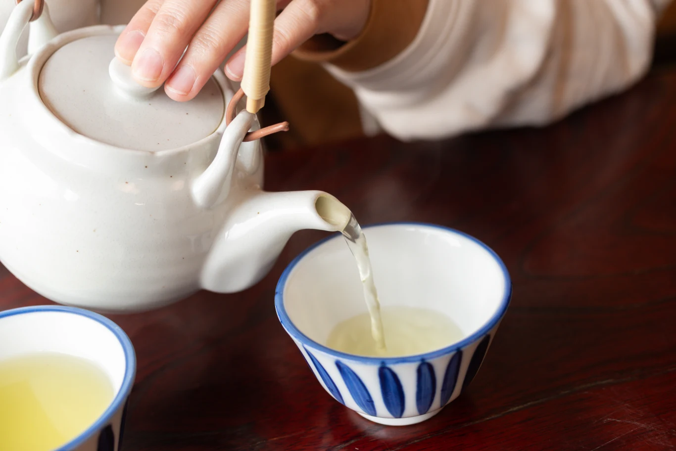 Ręka nalewająca gorący napój z białego dzbanka do filiżanki z charakterystycznym niebieskim wzorem na drewnianym stole.
