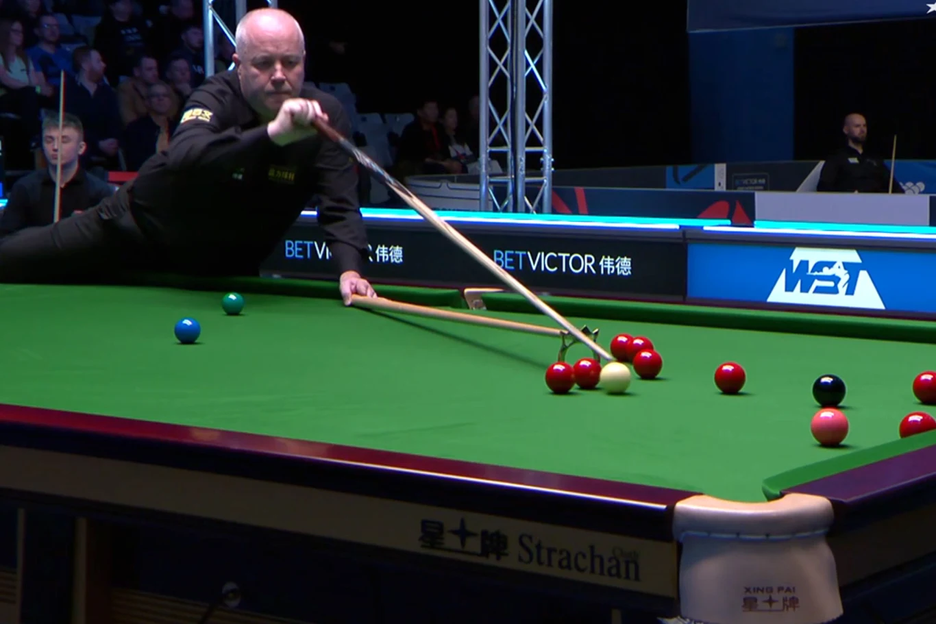 Mężczyzna w czarnym stroju wykonuje uderzenie kijem bilardowym podczas meczu snookera. Na stole widoczne są kolorowe bile, kilka osób obserwuje sytuację z trybun w tle. Silne oświetlenie skupia uwagę na graczu i stole.