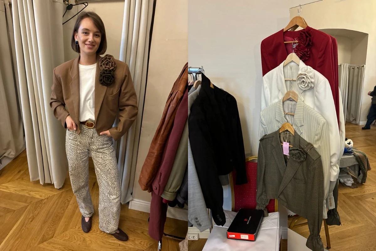 Młoda kobieta w beżowej marynarce i jasnych spodniach stoi przy zasłonce, obok zestaw wieszaków z różnokolorowymi marynarkami i swetrami ozdobionymi dużą broszką-kwiatem, widoczny drewniany parkiet i stół z pudełkiem.