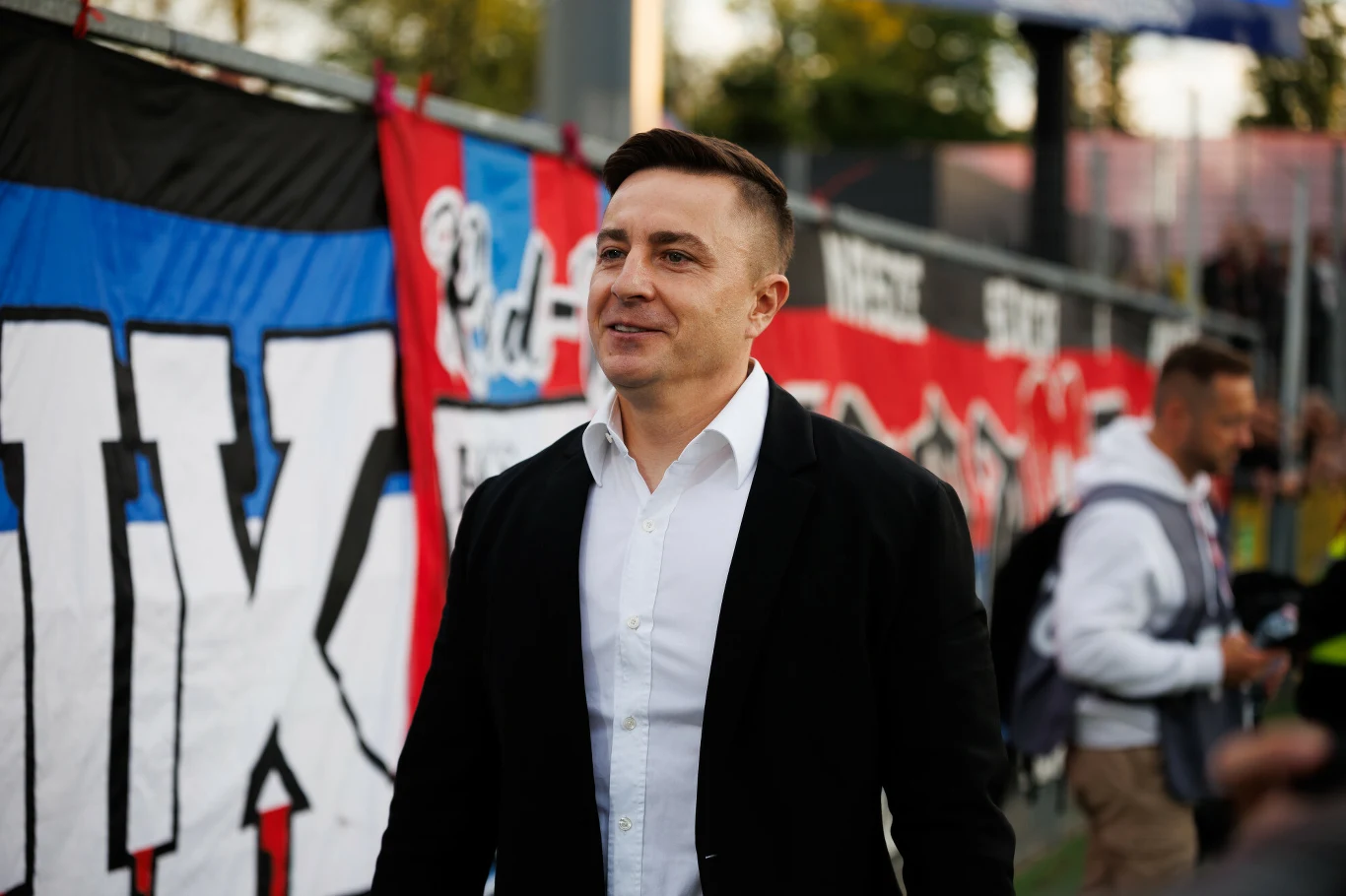 Michał Świerczewski Mężczyzna w marynarce i białej koszuli uśmiecha się, stojąc na tle barwnych transparentów sportowych, w tle widoczne osoby oraz elementy stadionu.