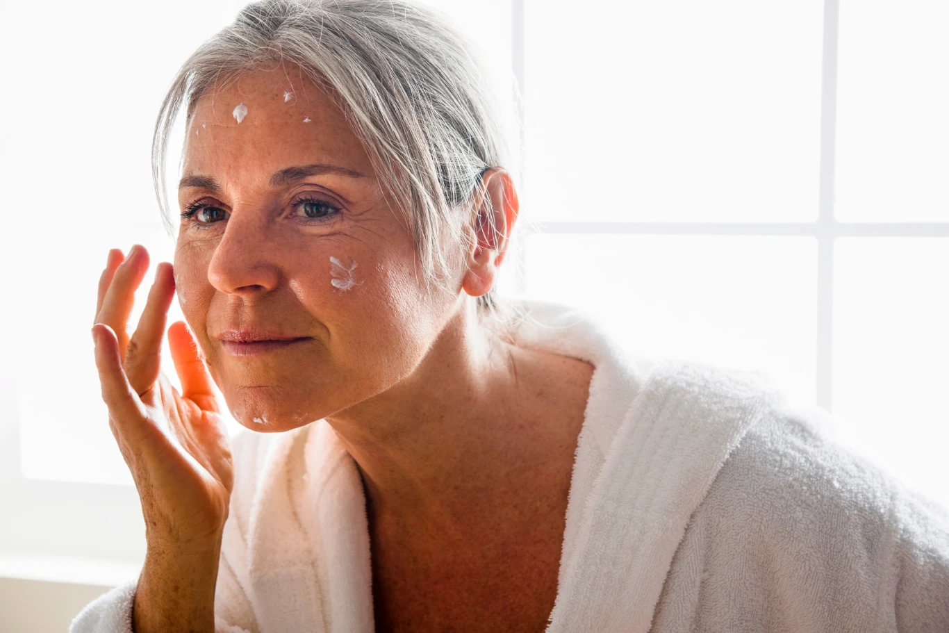 Starsza kobieta z siwymi włosami nakłada krem na twarz, ubrana w biały szlafrok, jasne światło w tle podkreśla naturalność sytuacji i codzienne dbanie o pielęgnację skóry.