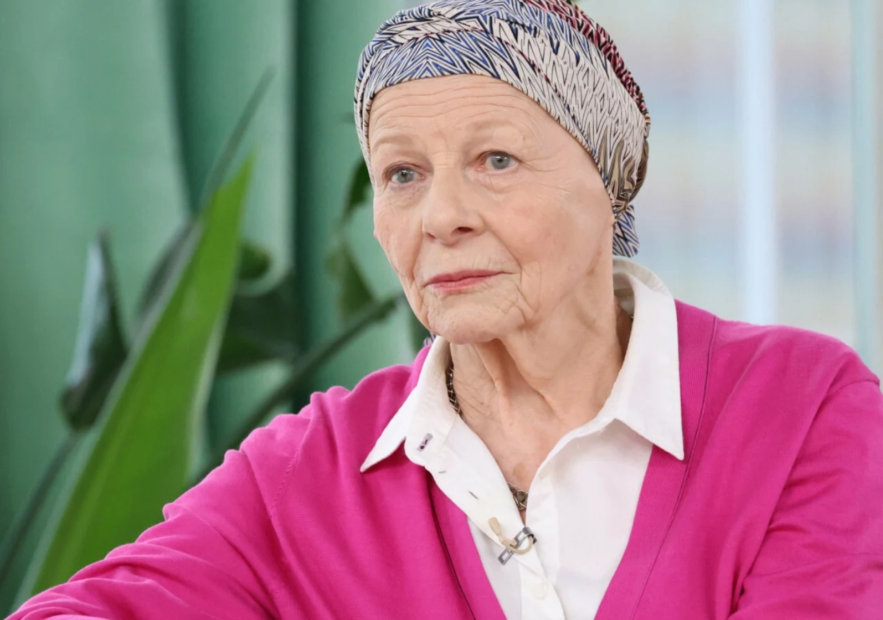 Anna Nehrebecka Starsza kobieta w apaszce na głowie i różowym swetrze siedzi we wnętrzu z zieloną rośliną w tle, ubrana elegancko i z zamyśloną miną.