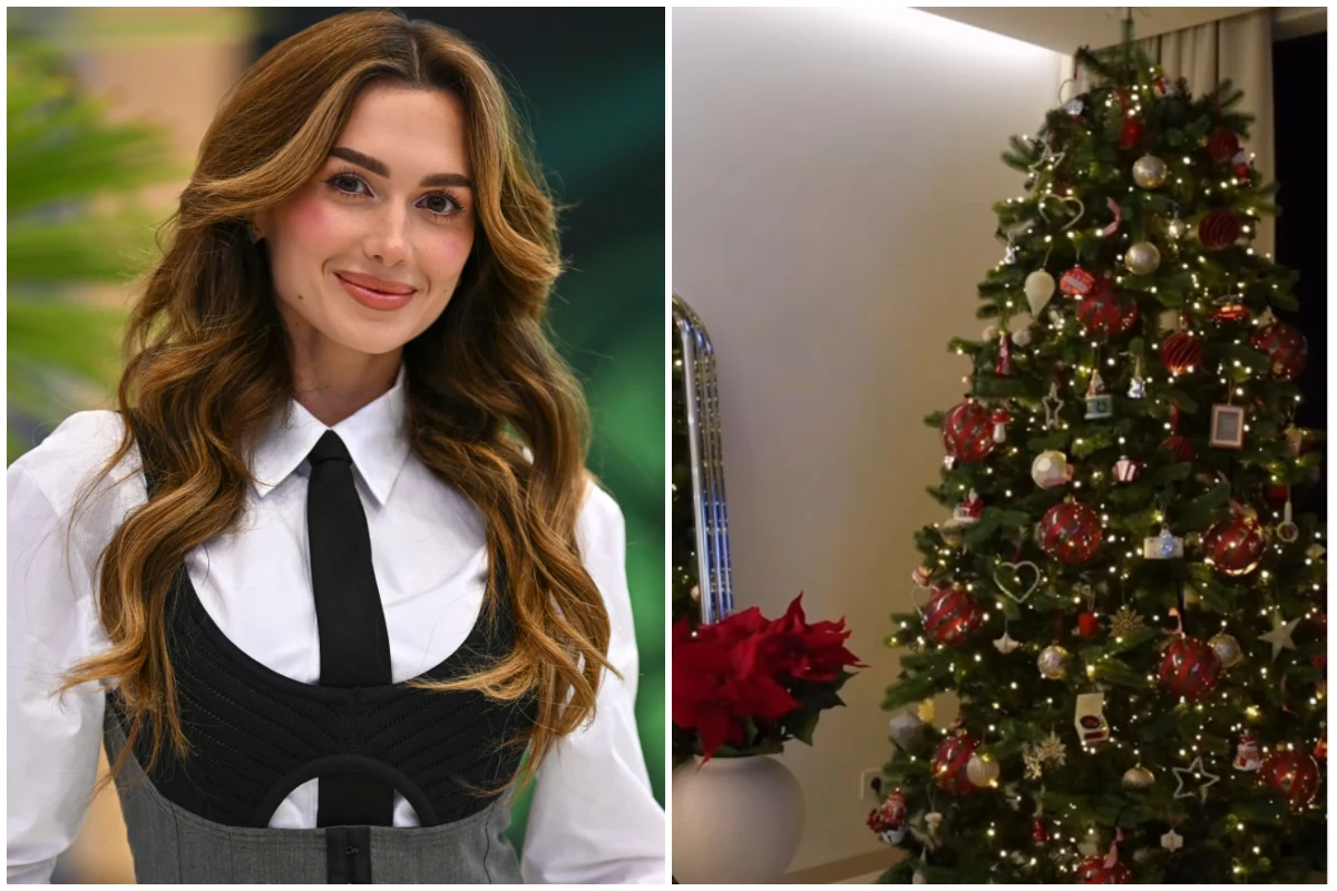 Uśmiechnięta kobieta o długich, falowanych włosach w eleganckim stroju obok bogato udekorowanej choinki z czerwonymi i złotymi bombkami oraz światełkami. Obok drzewka czerwone poinsecje w białej donicy.