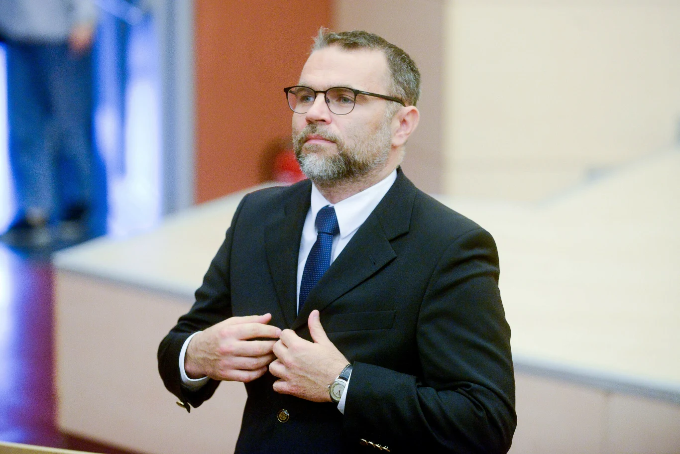 Jacek Bartosiak, publicysta zajmujacy sie geopolityką Mężczyzna w średnim wieku z brodą i okularami, ubrany w ciemny garnitur oraz krawat, stoi w pomieszczeniu o nowoczesnym wystroju, poprawiając marynarkę.