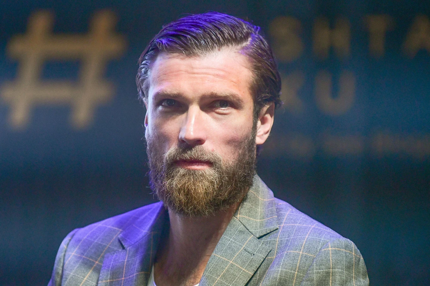 Andrzej Wrona Brodaty mężczyzna w eleganckiej marynarce, o poważnym wyrazie twarzy, na rozmytym tle z żółtymi napisami.