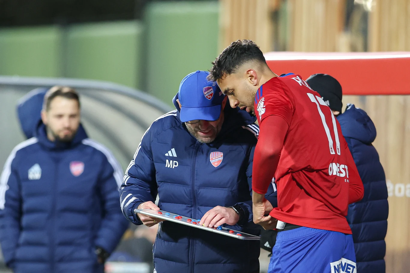 Trener drużyny piłkarskiej objaśnia taktykę jednemu z zawodników w czerwono-niebieskim stroju, korzystając z tablicy taktycznej, pozostali członkowie sztabu stoją w tle.