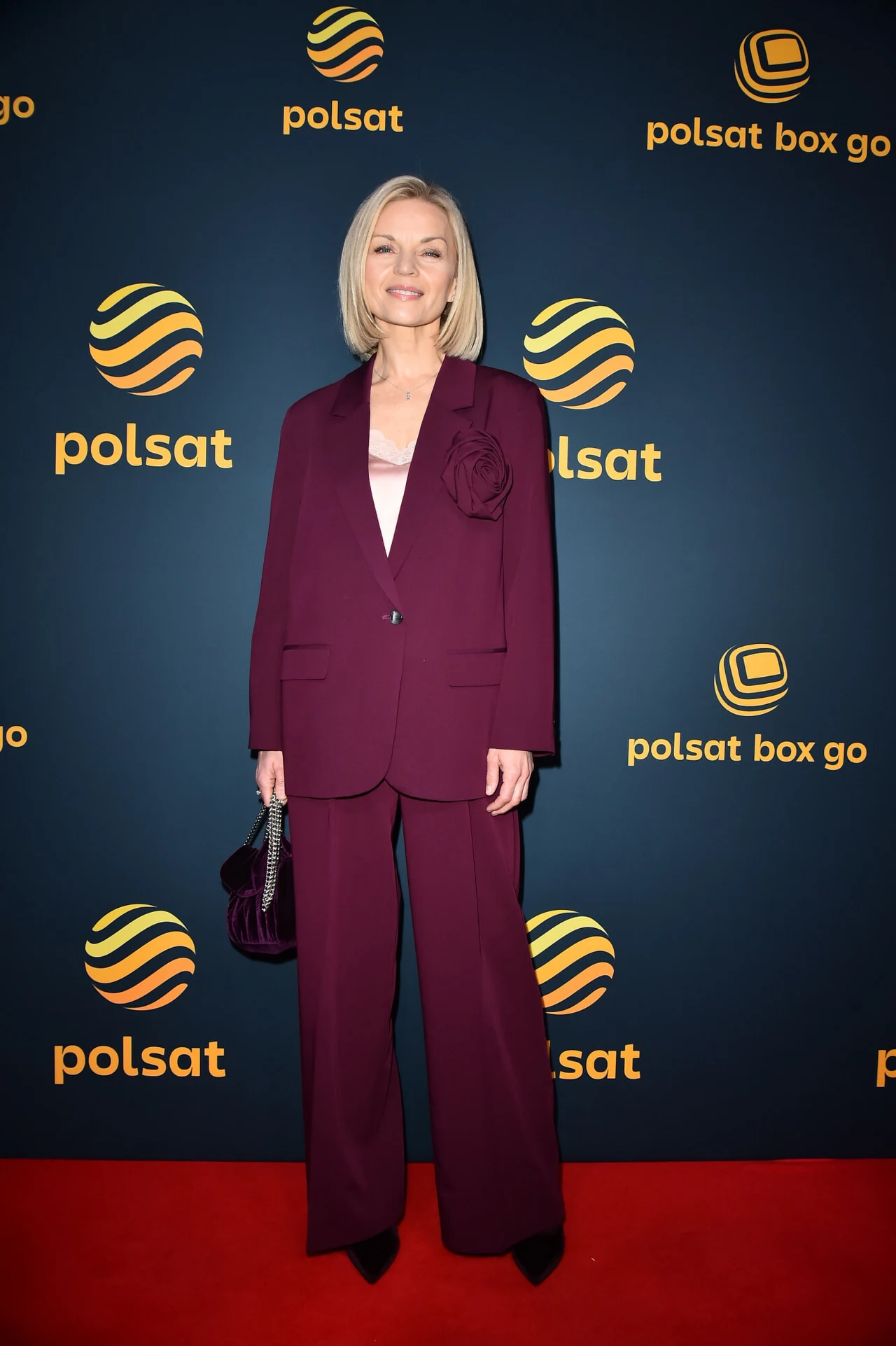 Małgorzata Foremniak zestawiła luźne spodnie z marynarką w modnym kolorze burgundowym. Zdobienie w postaci róży sprawiło, że stylizacja zyskała bardziej odświętny charakter Kobieta w eleganckim bordowym garniturze z kwiatową aplikacją na marynarce stoi na czerwonym dywanie, w tle ścianka z logotypami Polsat i Polsat Box Go.