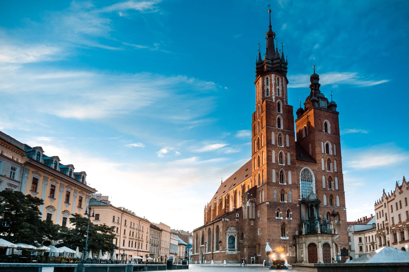 Kościół Mariacki o dwóch dominujących wieżach na tle błękitnego nieba, otoczony zabytkowymi kamienicami oraz otwartym placem rynku pełnym kawiarnianych ogródków.