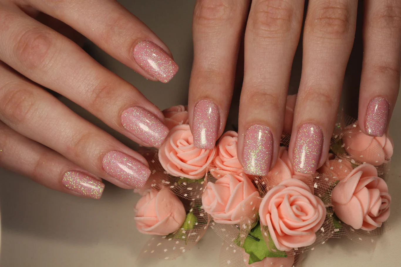 Dłonie z zadbanymi paznokciami pomalowanymi na różowo z połyskiem, ułożone na dekoracyjnych, sztucznych różach w pastelowym kolorze.
