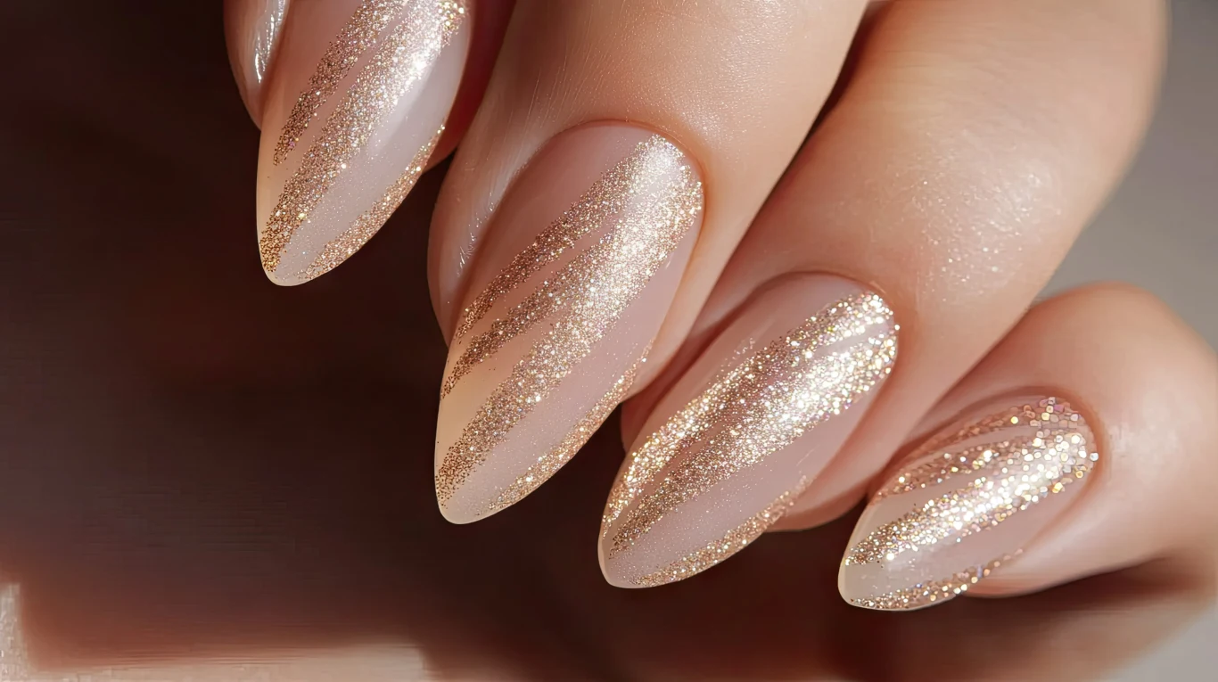 Zadbane dłonie z długimi paznokciami o migdałowym kształcie, pokryte jasnoróżowym lakierem z połyskującymi, złotymi brokatowymi zdobieniami.