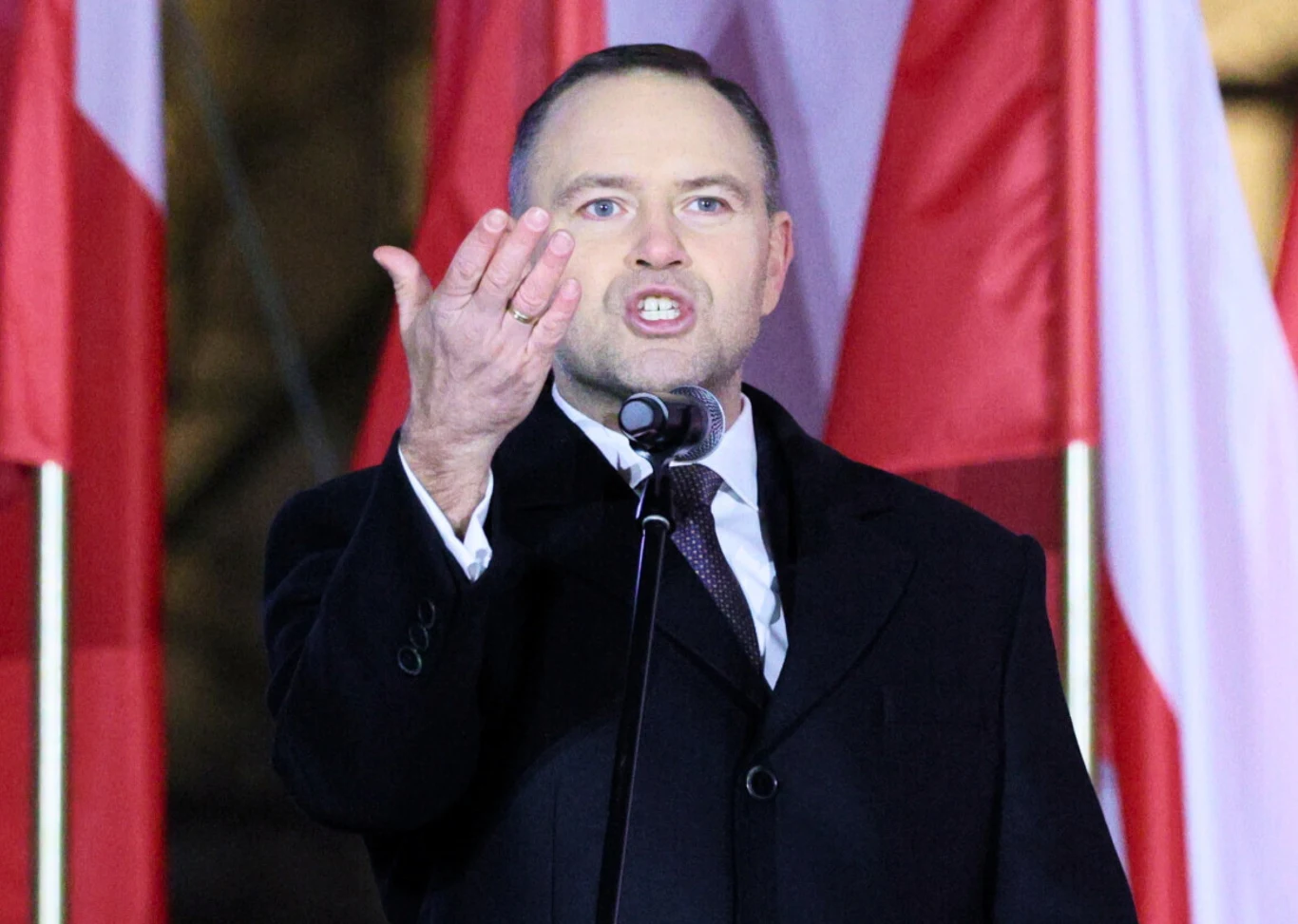 Mężczyzna w eleganckim płaszczu gestykuluje podczas przemówienia, stojąc przy mikrofonie na tle kilku polskich flag.
