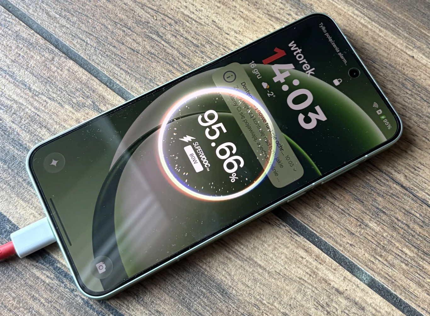 Smartfon leżący na drewnianej powierzchni podczas ładowania, wyświetlający na ekranie duży wskaźnik procentowy naładowania baterii oraz powiadomienia i aktualną godzinę.