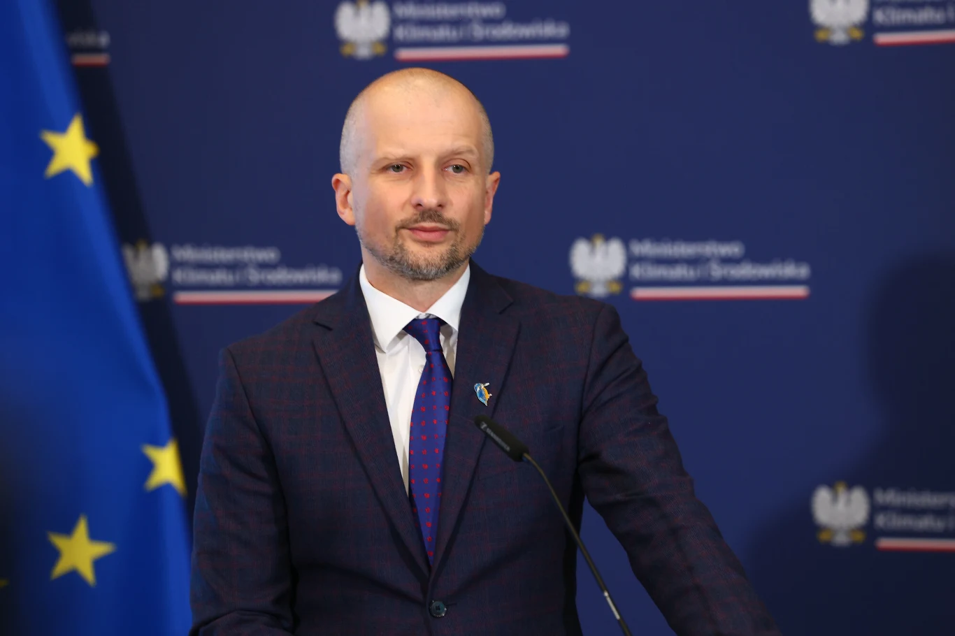 Głos w sprawie renaturyzacji doliny Nidy zabrał wiceminister Mikołaj Dorożała Mężczyzna w garniturze i krawacie stoi przy mikrofonie na tle oficjalnej ścianki z logotypami Ministerstwa Klimatu i Środowiska oraz flagą Unii Europejskiej.