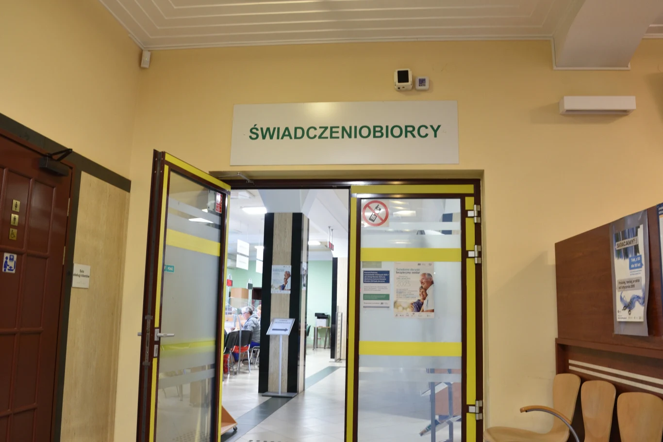 Emerytura wypłacana jest od dnia nabycia prawa do świadczenia. Wejście do sali urzędu lub instytucji z szyldem 'Świadczeniobiorcy', otwarte przeszklone drzwi, po lewej stronie widoczne krzesła i tablica informacyjna, w głębi widoczne stanowiska obsługi i osoby oczekujące.