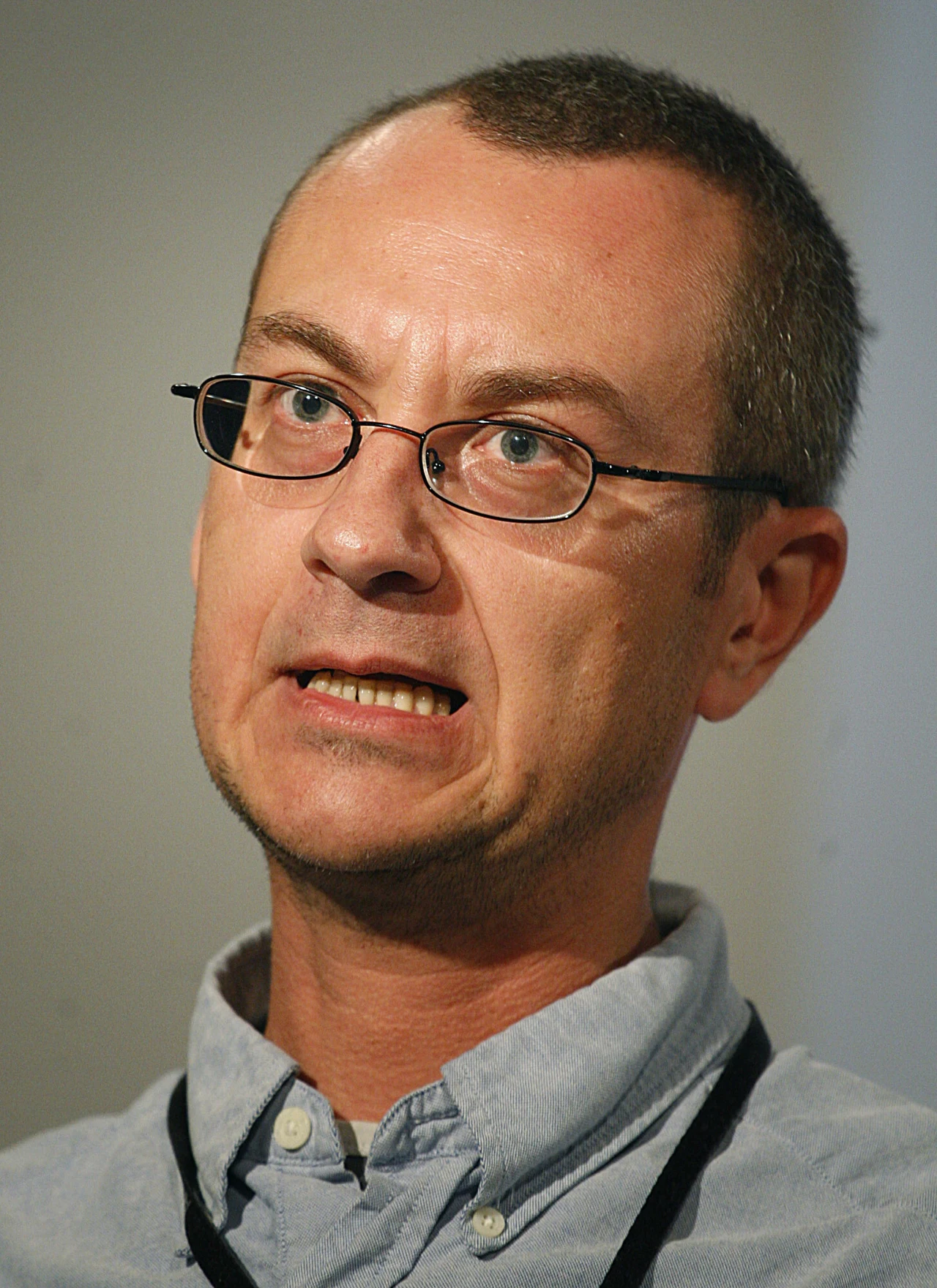 Mężczyzna w średnim wieku w okularach, z lekko zaciśniętymi zębami i napiętą mimiką twarzy, ubrany w jasną koszulę i ciemny sznurek na szyi, fotografowany na neutralnym tle.