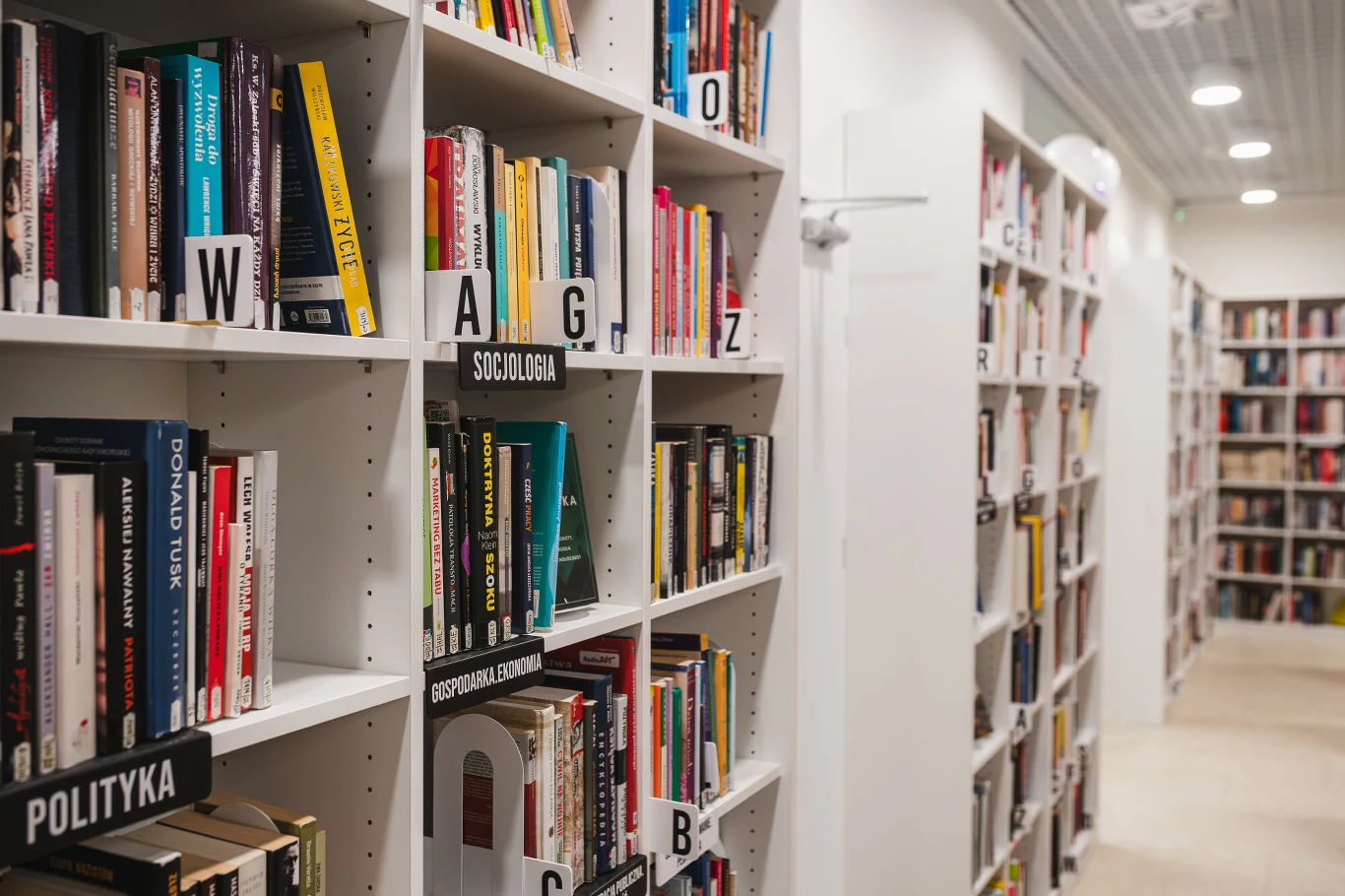 Półki biblioteczne wypełnione książkami, ułożonymi tematycznie według działów takich jak socjologia, polityka oraz literatura. Jasne wnętrze, białe regały oraz dobrze uporządkowany układ tworzą przestrzeń sprzyjającą nauce i czytaniu.