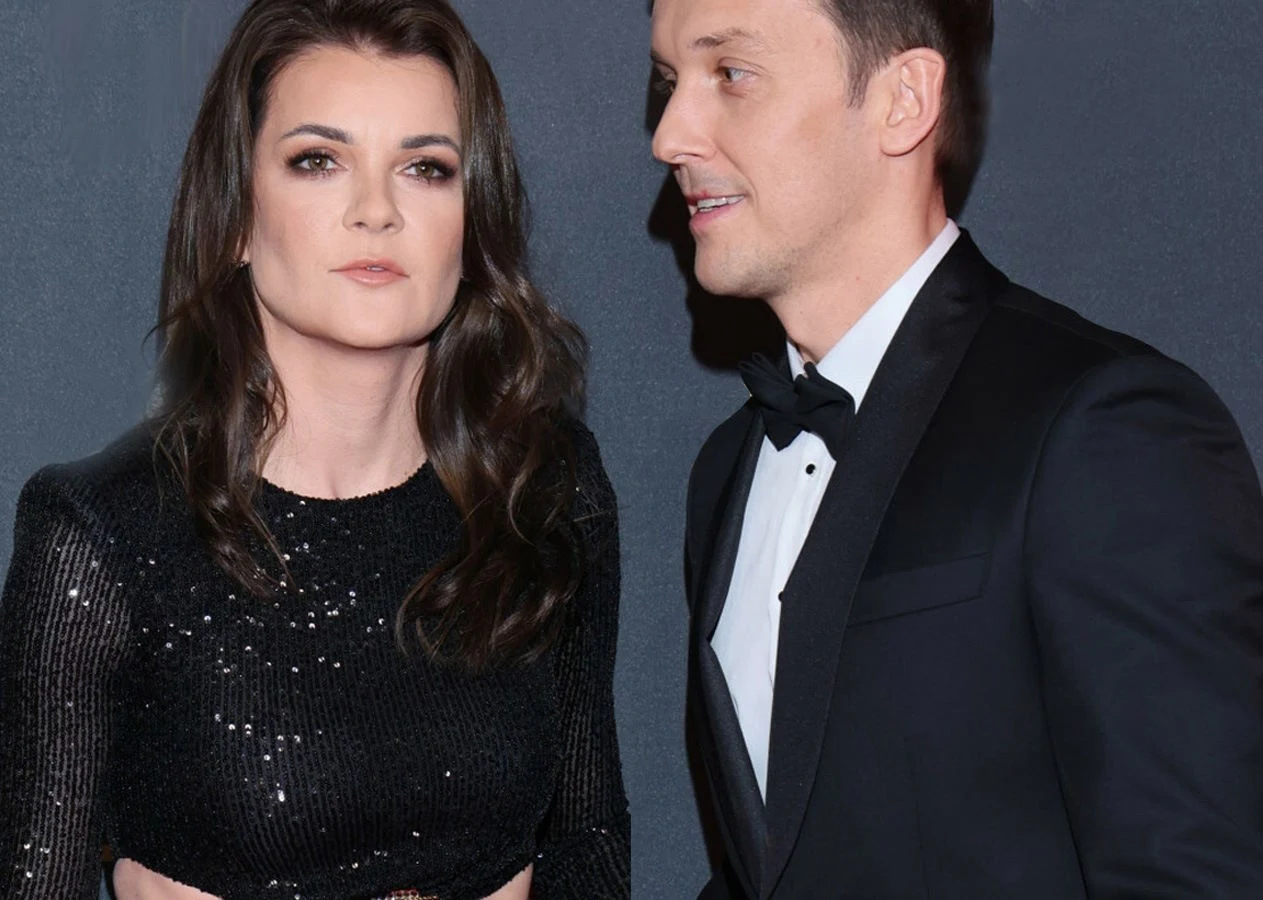 Agnieszka Radwańska i Dawid Celt Kobieta w czarnej, błyszczącej sukni oraz mężczyzna w eleganckim garniturze i muszce stoją razem na tle ciemnej ściany. Oboje mają poważny wyraz twarzy.